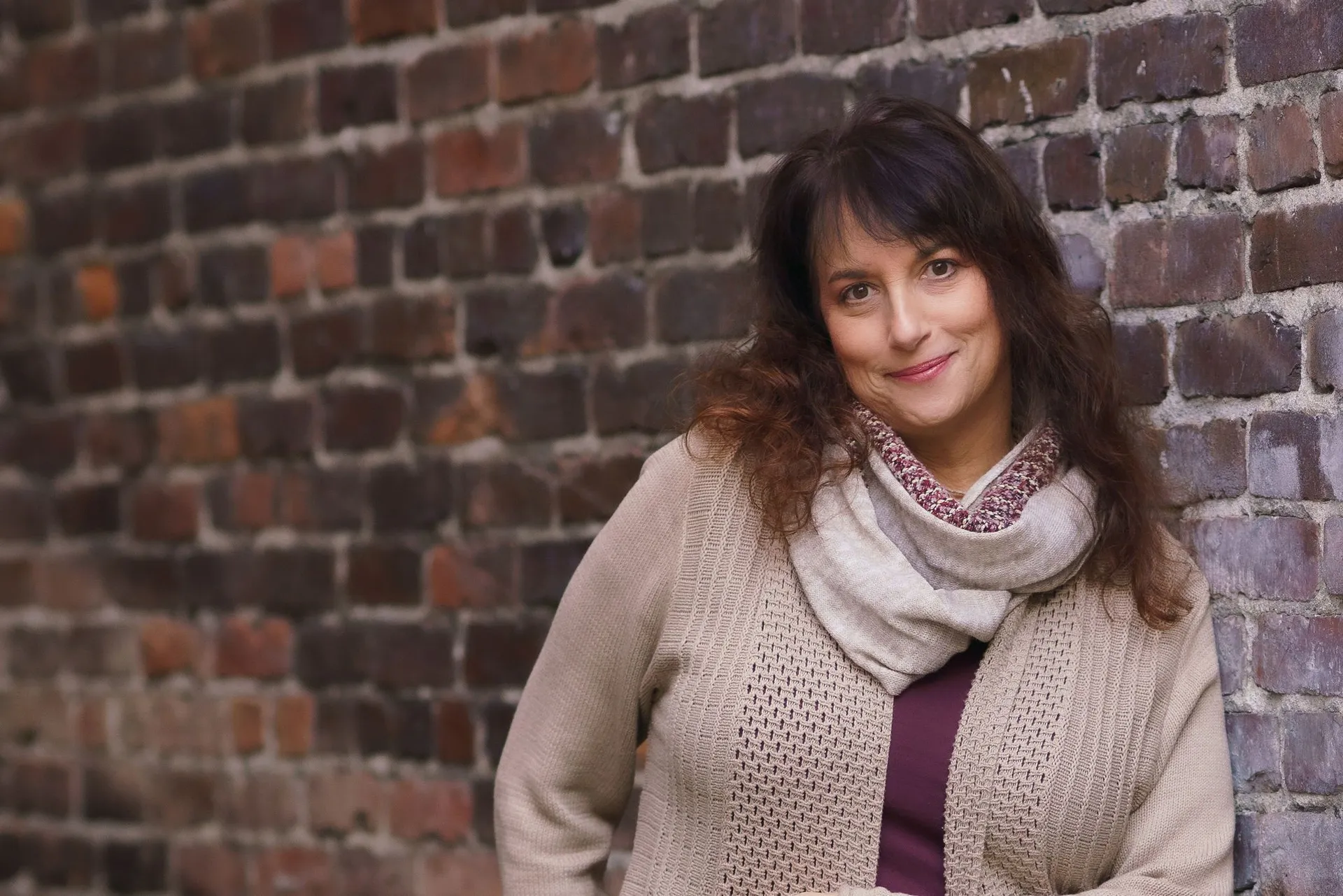 Issy Kleiman, MA, LMFT, AAMFT Approved Supervisor, looking at the camera beside a brick wall in a camel cardigan and scarf