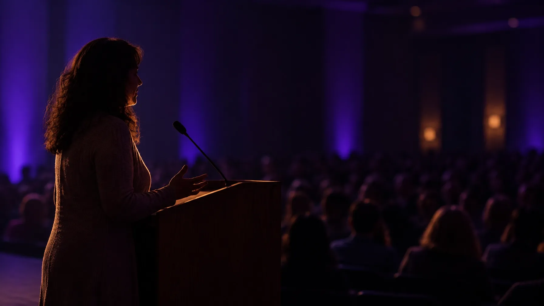 Speaker in profile addressing a large blurred audience from a wooden lectern under dim violet and indigo stage lighting